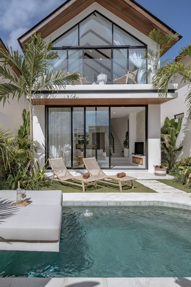 Modern two-story tropical villa with floor-to-ceiling glass windows, private swimming pool, sun loungers, balcony terrace, and lush palm landscaping under a clear blue sky.