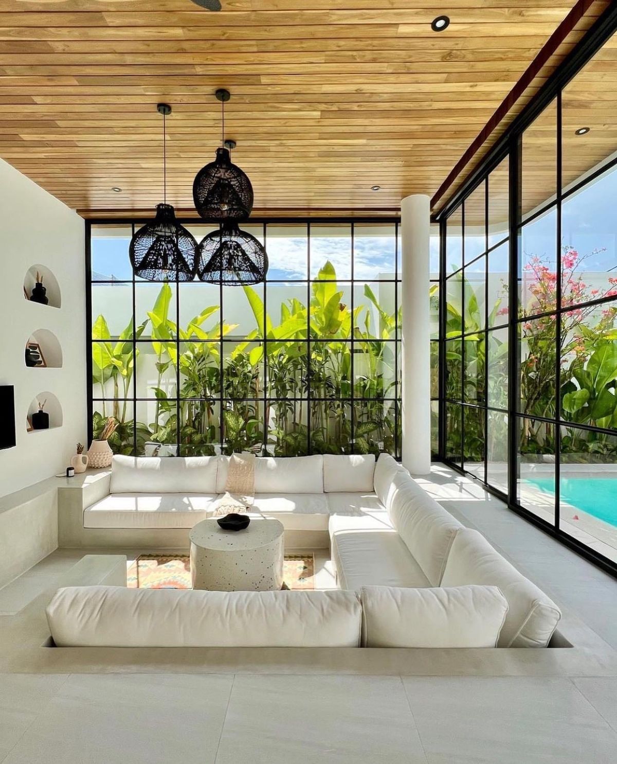 Modern tropical living space reflecting the concept of build villa in Bali, featuring floor-to-ceiling glass walls, wooden ceiling panels, sunken lounge seating, and a seamless connection to a private pool and lush garden.