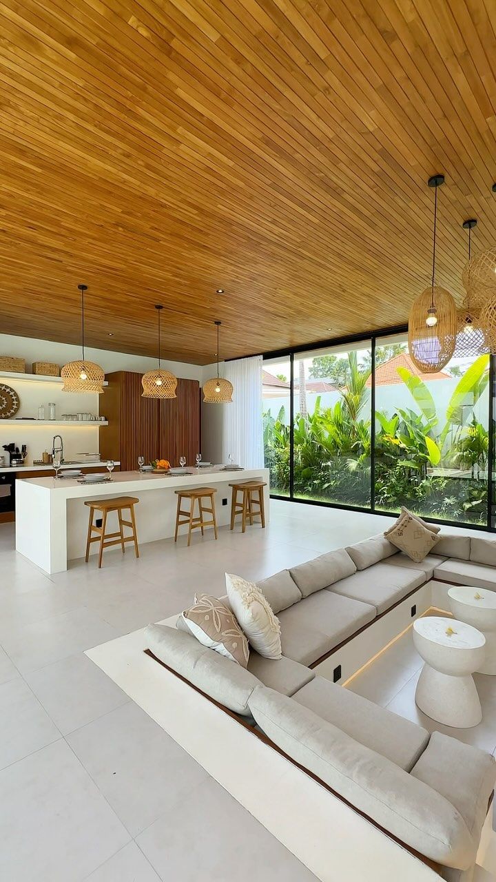 Modern tropical open-plan living room with a wooden ceiling, white kitchen island and bar stools, woven pendant lights, sunken sofa seating area, and large sliding glass doors overlooking a lush garden.