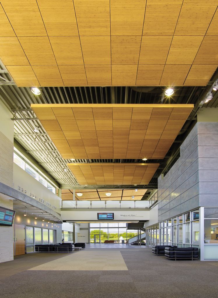 Modern commercial lobby featuring suspended wooden drop ceiling panels with acoustic perforation, creating a layered ceiling design beneath exposed structural elements and industrial lighting.